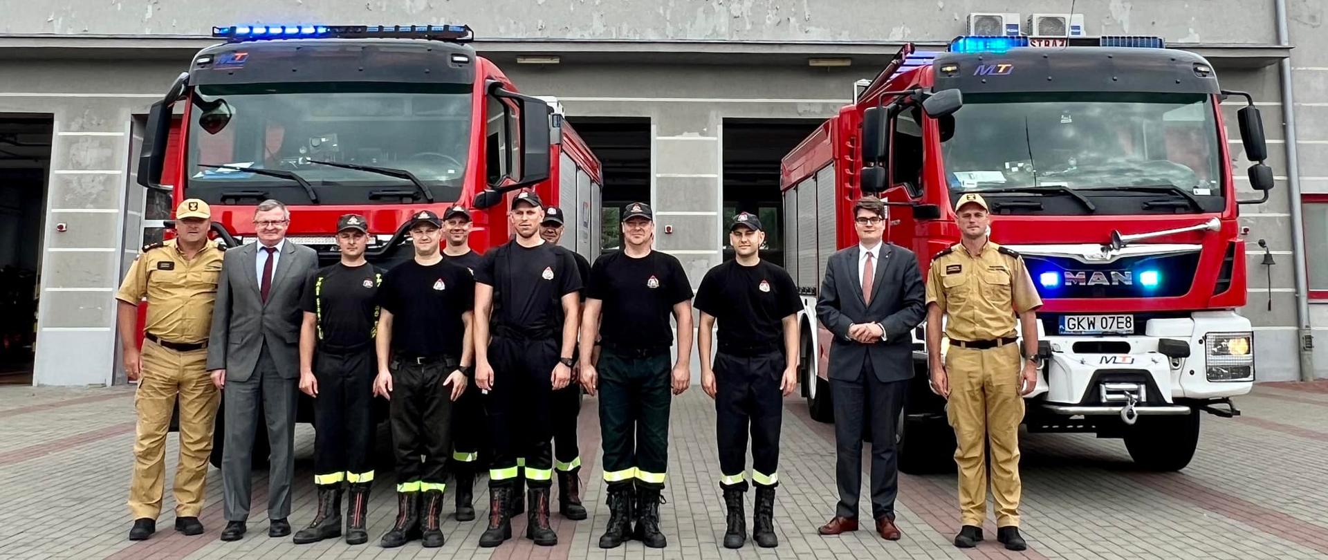 Komendanci i strażacy komendy PSP w Kwidzynie wraz z Wicewojewodą Pomorskim oraz Posłem na Sejm pozują do zdjęcia na tle samochodów pożarniczych.