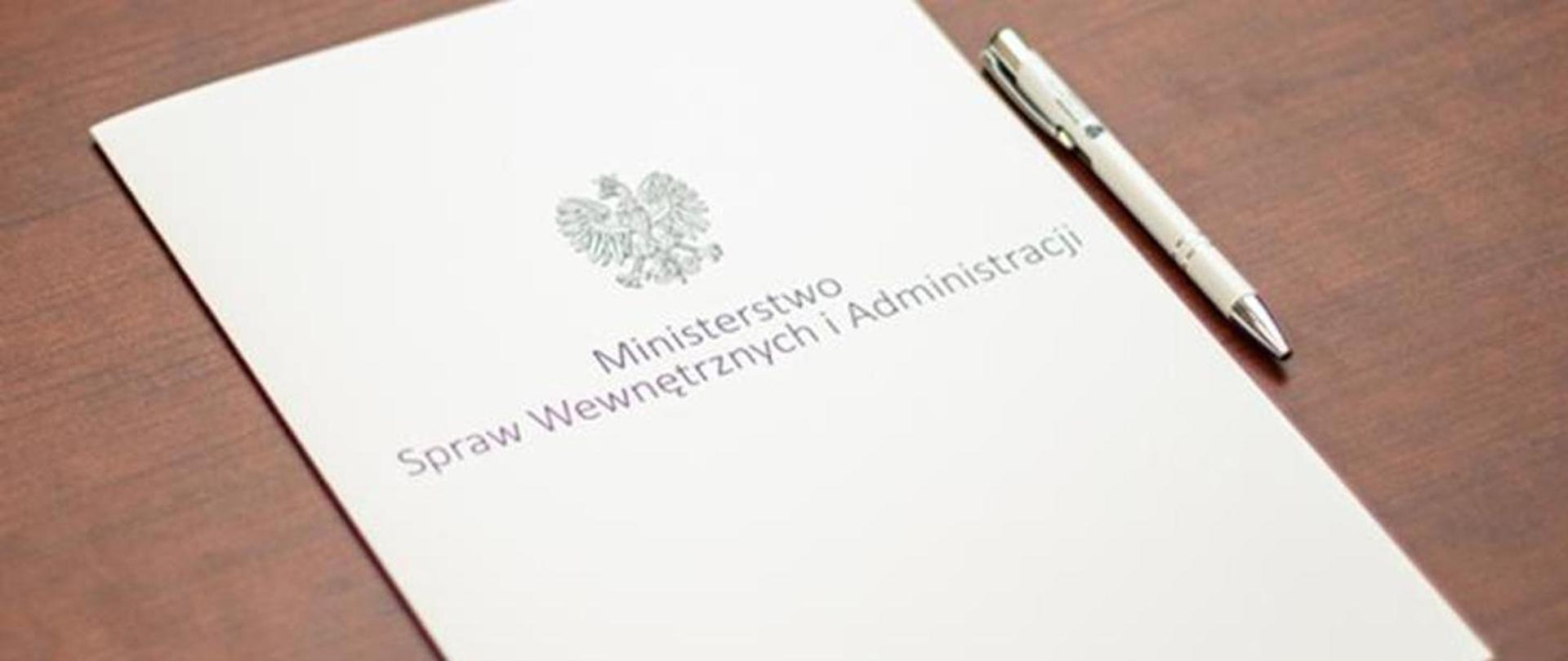 Biała_teczka_z_napisem_Ministerstwo_Spraw_Wewnętrznych_i_Administracji_lezy_brązowym_stole_Obok_teczki_leży_biały_długopis