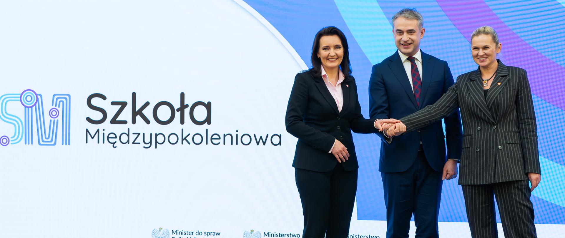 Startuje konkurs „Szkoła międzypokoleniowa” skierowany do szkół podstawowych i ponadpodstawowych podłączonych do Ogólnopolskiej Sieci Edukacyjnej. Konkurs ogłoszony na konferencji prasowej w KPRM. Na zdjęciu z prawej strony ministrowie podający sobie ręce. po lewej czarny napis szkoła międzypokoleniowa. 