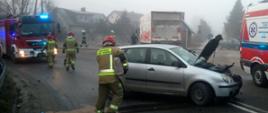 Na zdjęciu znajduje się uszkodzony samochód osobowy marki VW Polo z podniesioną maską. Przed samochodem znajduje się strażak likwidujący wyciek płynów eksploatacyjnych. W tle widać samochód strażacki, Karetkę Pogotowia Ratunkowego oraz strażaków jak również samochód ciężarowy stojący po przeciwnej stronie jezdni
