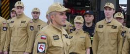 Zdjęcie przedstawia awansowanego na stanowisko dowódcy zmiany asp. sztab. Arkadiusza Kopczyńskiego w mundurze służbowym. Za nim w dwuszeregu stoją funkcjonariusze Komendy Powiatowej PSP w Brodnicy.