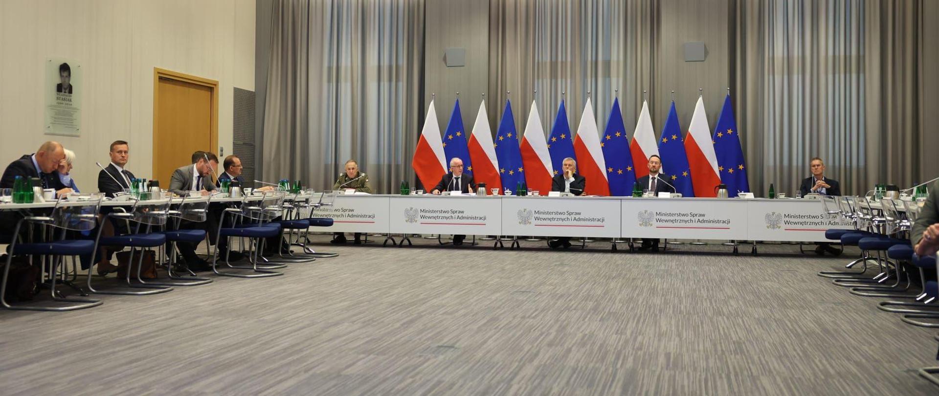 Ministrowie siedzą przy środkowym stole. Wojewodowie siedzą przy stołach po lewej i prawej stronie. Za ministrami widać flagi Polski i Unii Europejskiej, każdej jest po 6.