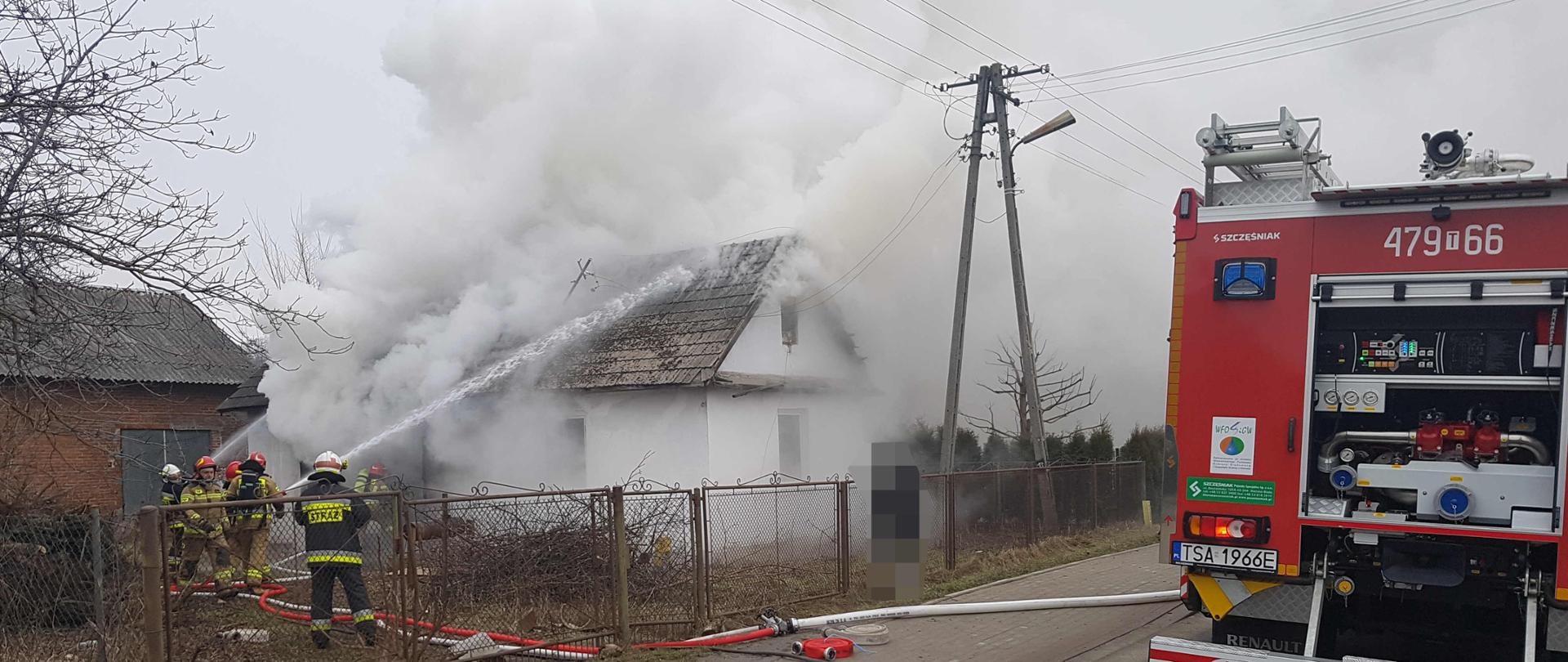 zdjęcie zrobione w dzień. Na zdjęciu widać palący się budynek mieszkalny i dużo dymu. Zdjęcie przedstawia strażaków, którzy rozwijają węże i prowadzą akcję gaśniczą budynku. W dalszej części zdjęcia widać tył samochodu strażackiego. 