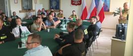 Na zdjęciu widoczna sala szkoleniowa KM PSP. Widzimy również przedstawicieli władz samorządowych i Komendy Miejskiej PSP w Tarnowie. W tle flagi Polski oraz godło. Z lewej strony zdjęcia widać uczestników spotkania.