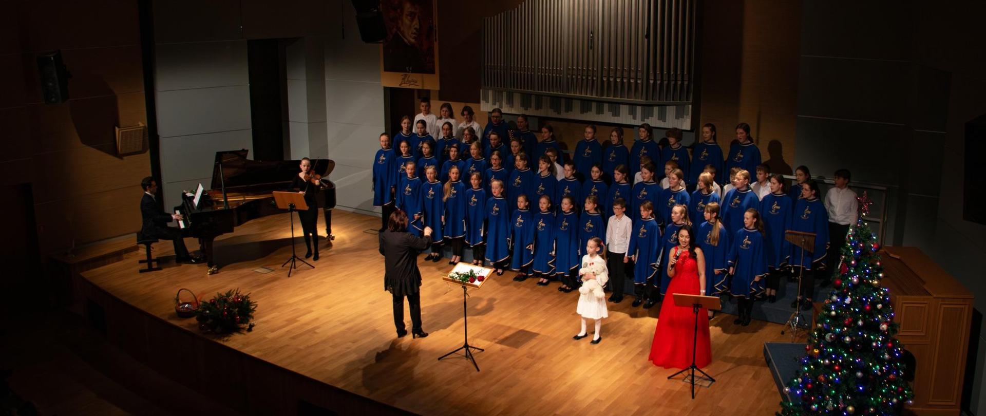 Zdjęcie przedstawiające występujący na scenie sali koncertowej szkoły chór, osobę prowadzącą chór, pianistę akompaniującego na fortepianie oraz uczennicę grającą na skrzypcach, kilkuletnią ubraną na biało uczennicę śpiewającą do mikrofonu, braną na czerwono solistkę - studentkę AM wydziału wokalnego. Po prawej stronie sceny widoczna ozdobiona świąteczna choinka.