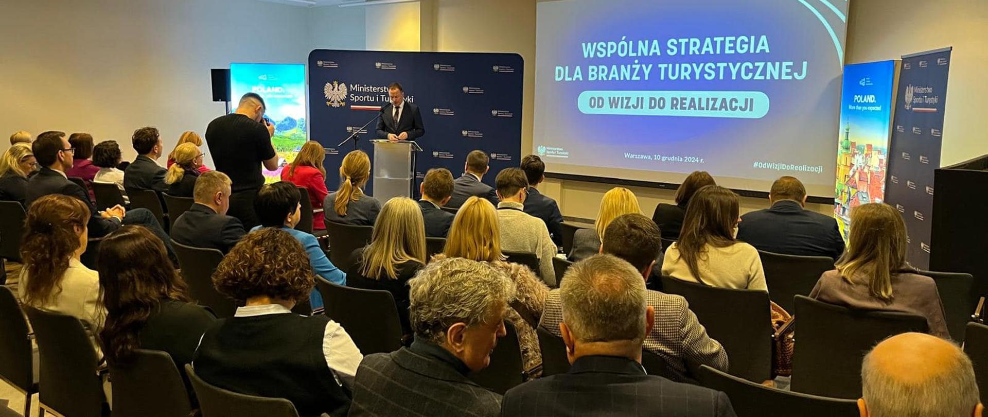 Zdjęcie z konferencji pn. „Wspólna strategia dla branży turystycznej: od wizji do realizacji”. Na pierwszym planie goście konferencji, w tle przemawiający Sekretarz Stanu w Ministerstwie Sportu i Turystyki Piotr Borys.