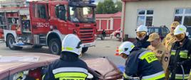 na zdjęciu strażacy w trakcie wykonywania dostępu do osoby poszkodowanej
w trakcie założenia egzaminacyjnego, w tle pojazd pożarniczy