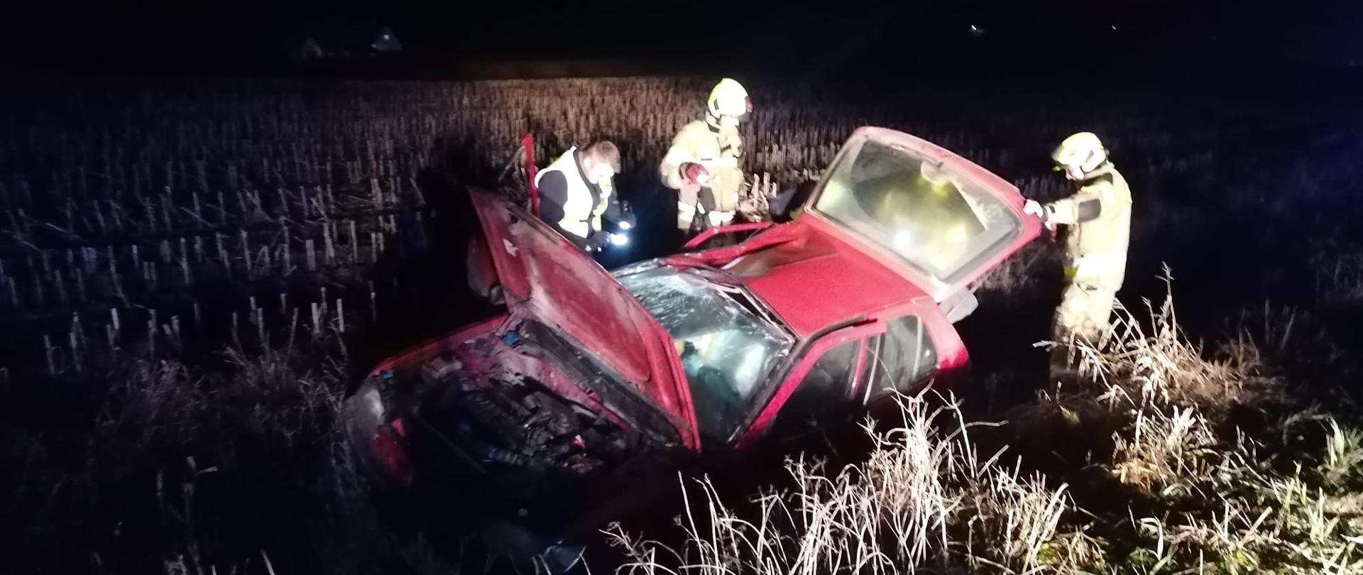 Zdjęcie zrobione nocą. Poza drogą w rowie stoi czerwone auto osobowe, uszkodzone, przy nim strażacy.