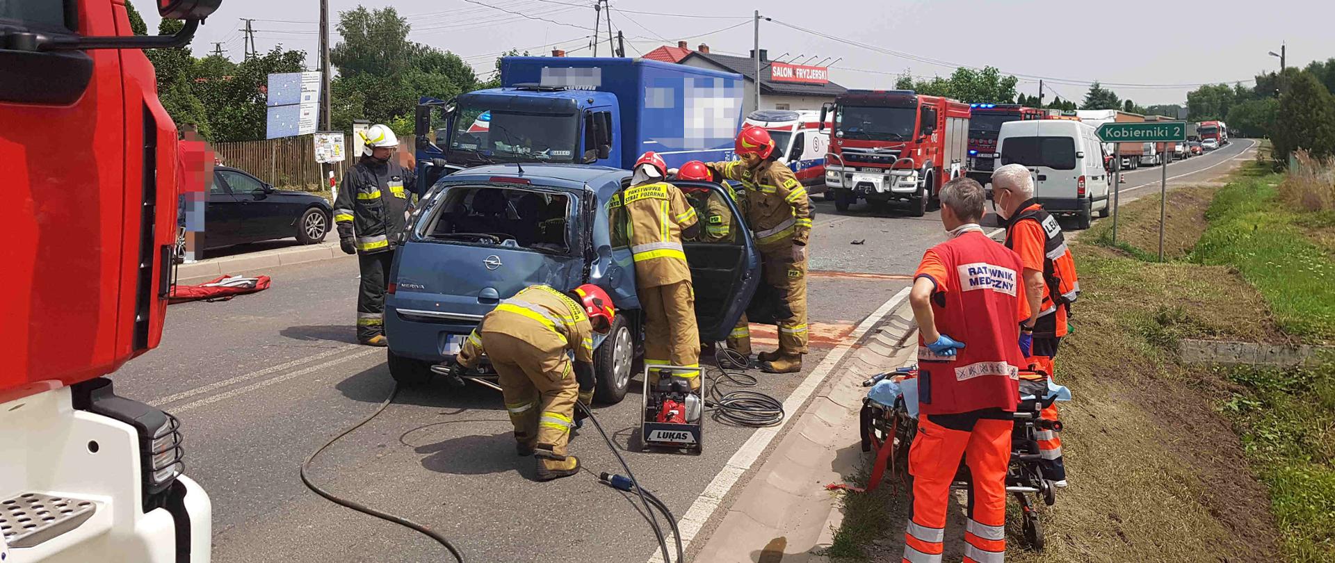 Zdjęcie zrobione w dzień. Zdjęcie przedstawia strażaków którzy robią dostęp do osoby poszkodowanej sprzętem hydraulicznym w aucie osobowym. W oddali widać ratowników medycznych oczekujących na przekazanie osoby poszkodowanej 