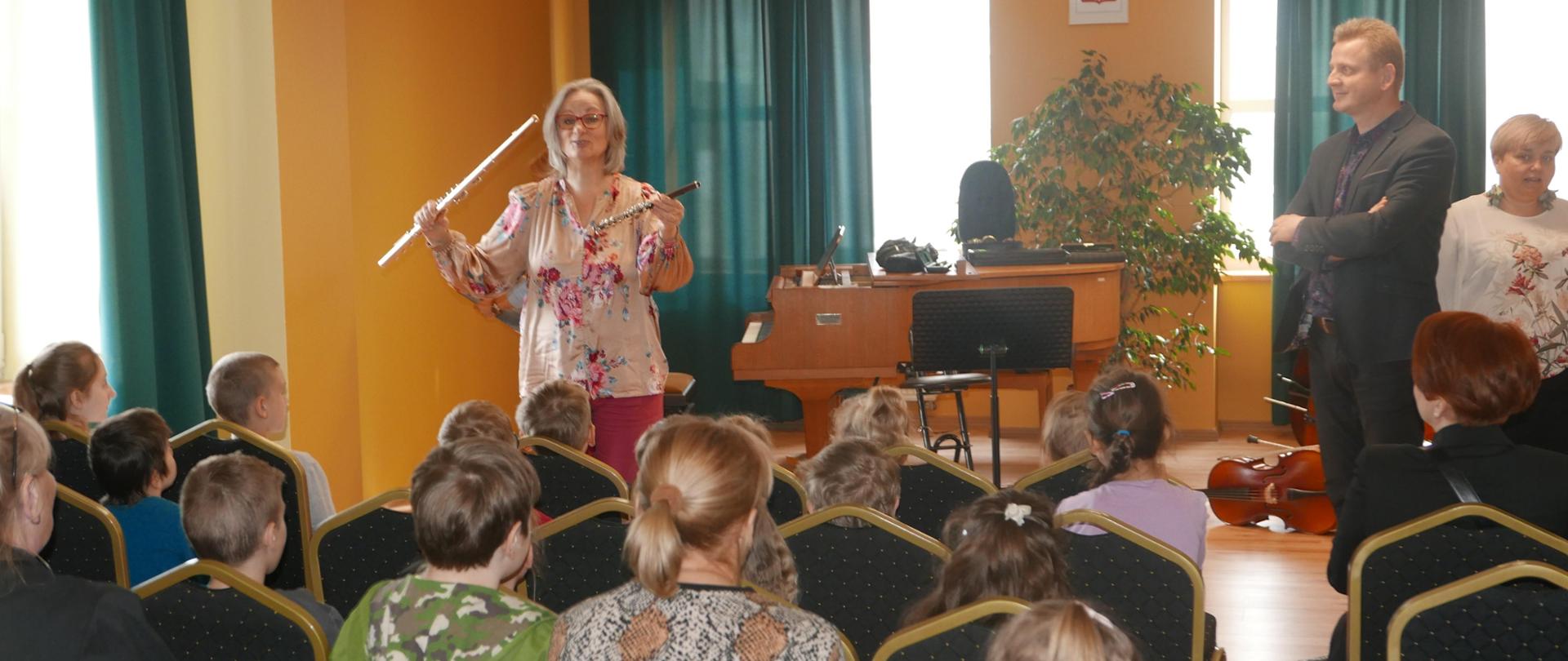 Widzimy widownię złożoną z przedszkolaków patrzących na to, co się dzieje na scenie. A tu widzimy panią prezentującą dwa flety poprzeczne, duży i picolo.