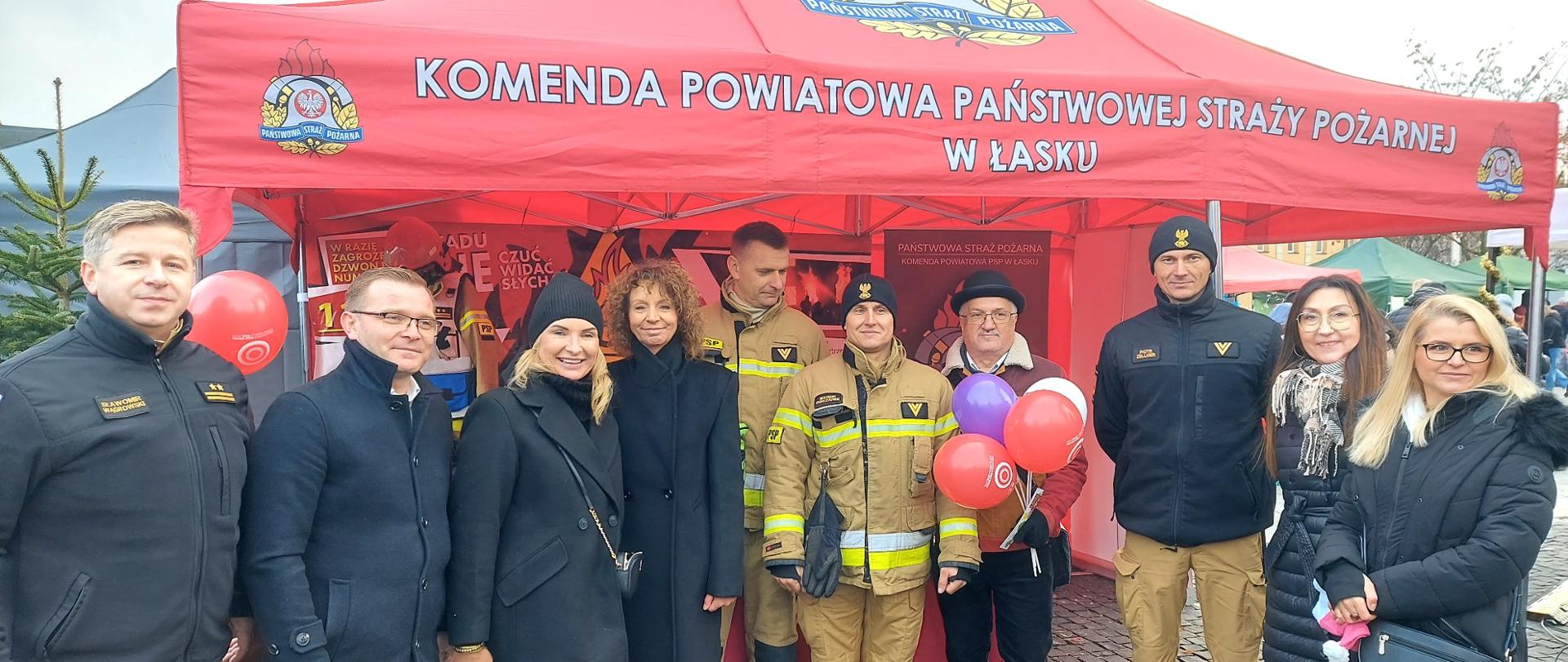 Na zdjęciu widać grupę ludzi stojących przed czerwonym namiotem z napisem: „KOMENDA POWIATOWA PAŃSTWOWEJ STRAŻY POŻARNEJ W ŁASKU”. Namiot jest ozdobiony logotypami PSP (Państwowej Straży Pożarnej). Na pierwszym planie stoi kilkoro strażaków w mundurach służbowych i ubraniach ochronnych, a także osoby w cywilnych strojach, w tym kobiety i mężczyźni, uśmiechający się do zdjęcia. W tle widać tablice informacyjne na temat działań straży pożarnej oraz dekoracje nawiązujące do świątecznego charakteru wydarzenia. Niektórzy trzymają balony w kolorach czerwonym i fioletowym, co dodaje atmosfery radości i zabawy.
Zdjęcie zostało wykonane podczas Jarmarku Bożonarodzeniowego, w ramach którego Komenda Powiatowa PSP w Łasku zorganizowała stoisko, aby promować bezpieczeństwo i edukować mieszkańców na temat działań strażackich. W tle można zauważyć inne stragany i choinkę, co podkreśla świąteczny klimat wydarzenia.