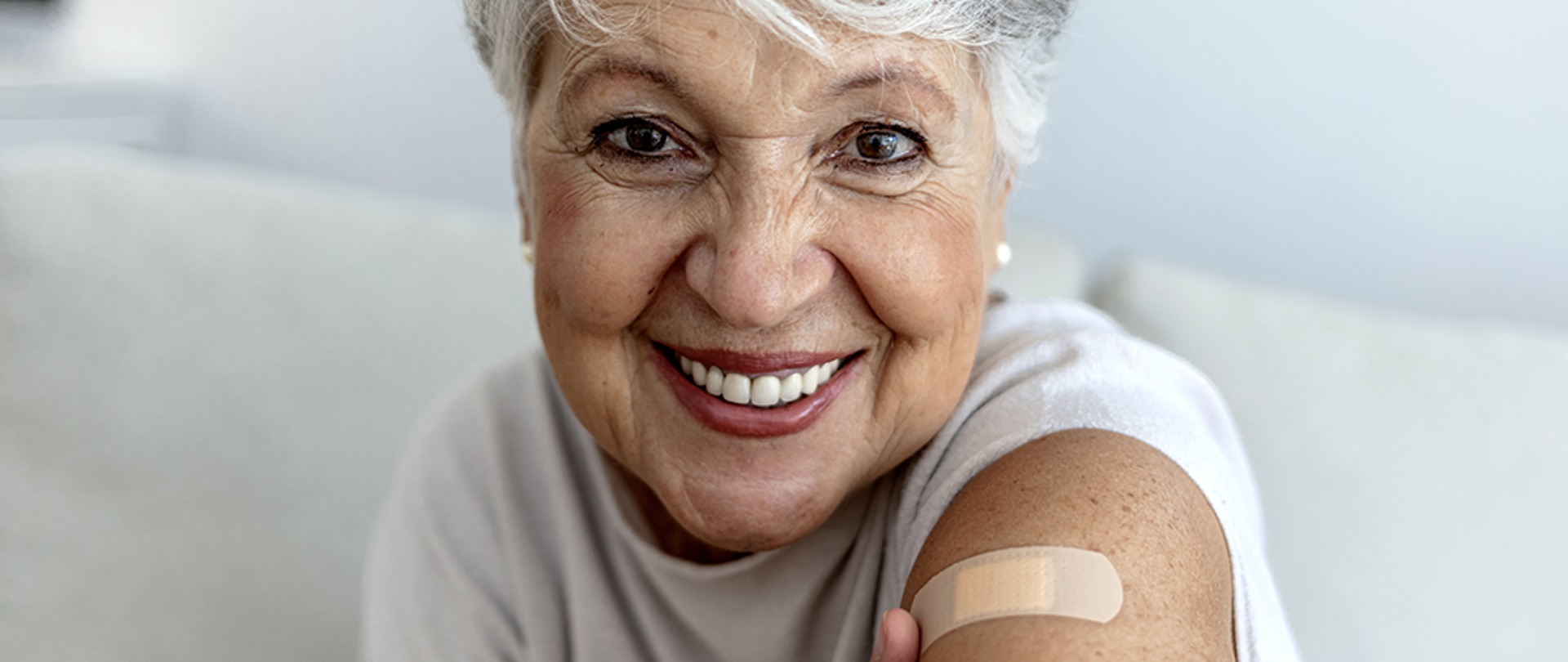 Na zdjęciu widoczna jest ok. 60-letnia kobieta. Jest uśmiechnięta, patrzy wprost na odbiorcę. Na jej ramieniu widoczny jest plaster, który został przyklejony tuż po zaczepieniu przeciw COVID-19.
