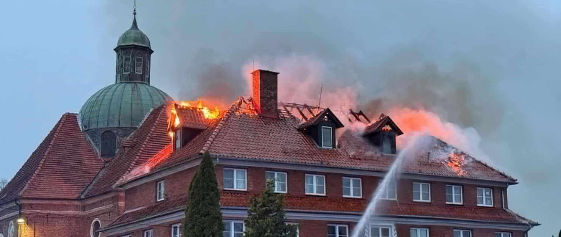 Płonie dach dużego budynku z cegły. Widać płomienie. Z lewej strony widać wieże kościoła. Strażacy leją wodę.