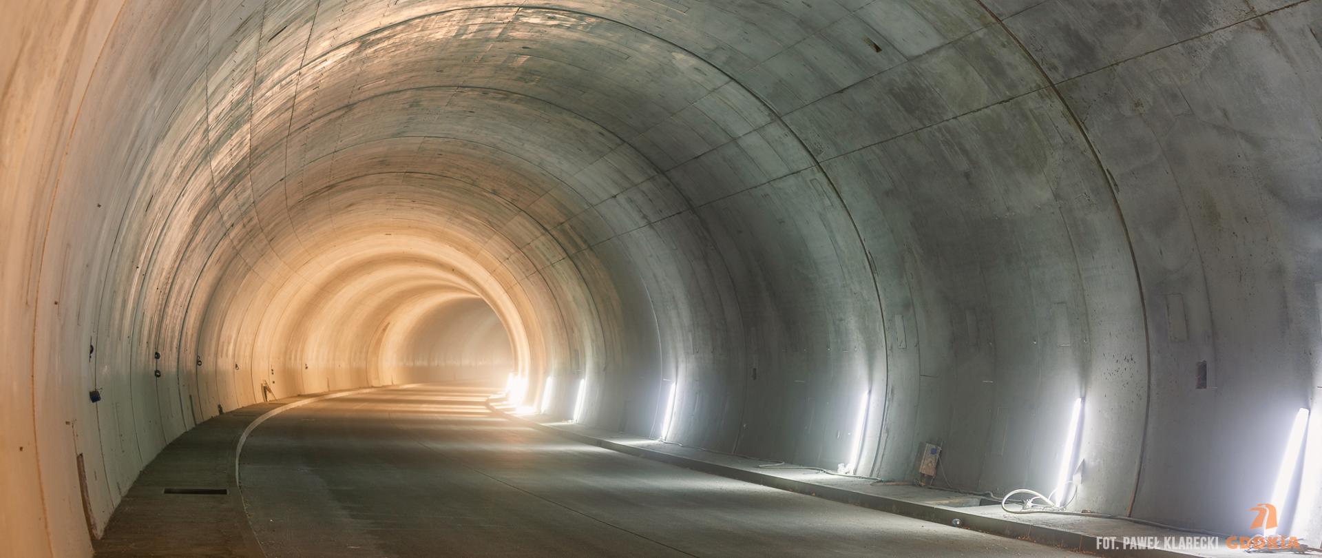 Zdjęcie przedstawia wnętrze tunelu drogowego. Tunel jest oświetlony jasnym światłem, które tworzy efektowne refleksy na jego betonowych ścianach. Ściany tunelu są gładkie i mają lekko zaokrąglony kształt. Na podłodze tunelu widać asfaltową nawierzchnię drogi. W tunelu nie ma żadnych pojazdów ani ludzi, co sprawia, że wygląda on na pusty i spokojny. 