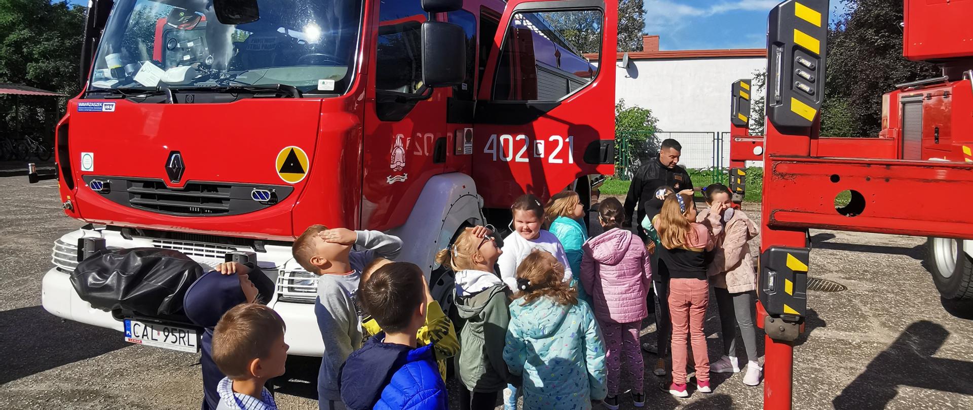 Na zdjęciu widać grupę uczniów klasy pierwszej ze Szkoły Podstawowej nr 3 w Ciechocinku, biorących udział w pokazie sprzętu strażackiego. Dzieci stoją w kolejce do wejścia do pojazdu ratowniczo gaśniczego. W tle widać samochód pożarniczy stojący na parkingu szkolnym.