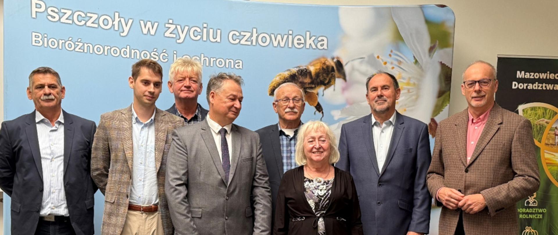 Na zdjęciu widać grupę ośmiu osób, które stoją na tle baneru z napisem „Pszczoły w życiu człowieka. Bioróżnorodność i ochrona”