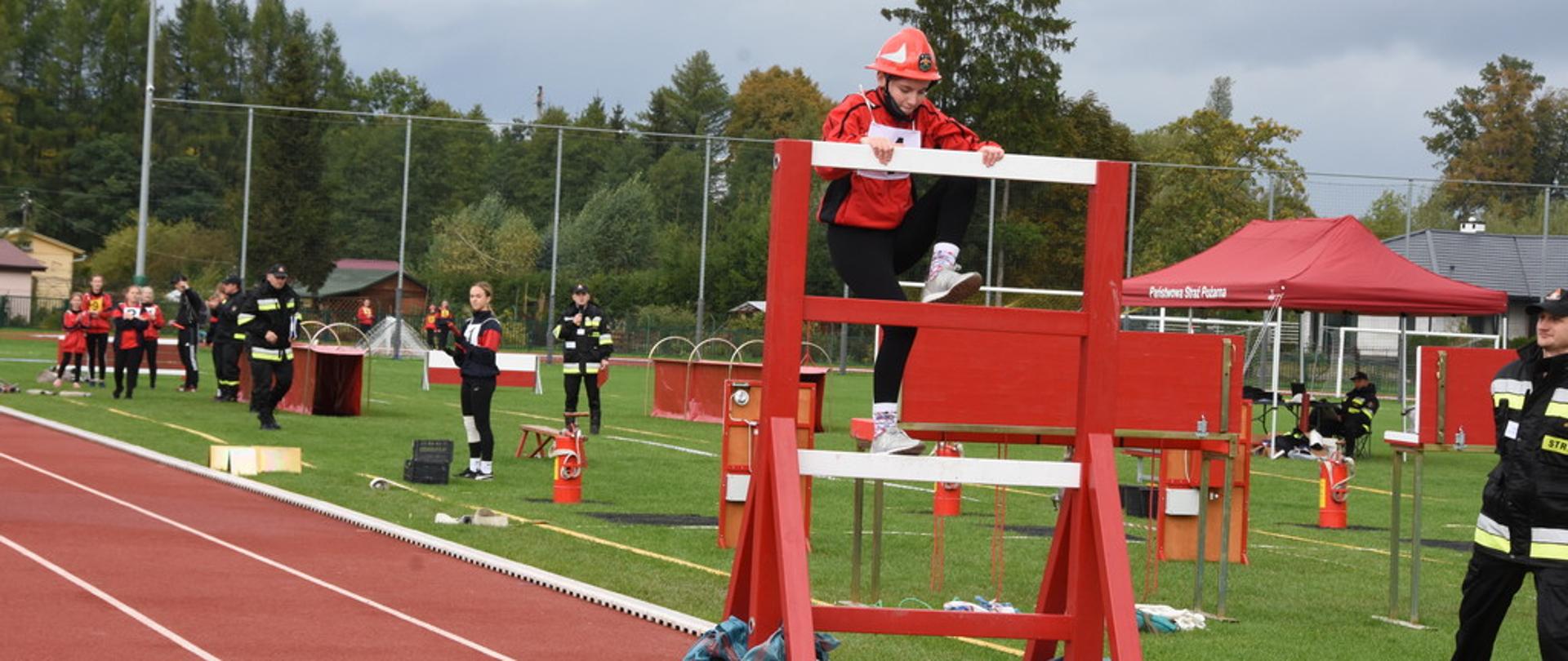 Na zdjęciu widoczny zawodnik Młodzieżowej Drużyny Pożarniczej pokonujący ścianę drabiniastą podczas zawodów sportowo – pożarniczych. W tle sędziowie oraz inna drużyna przygotowująca się do startu.
