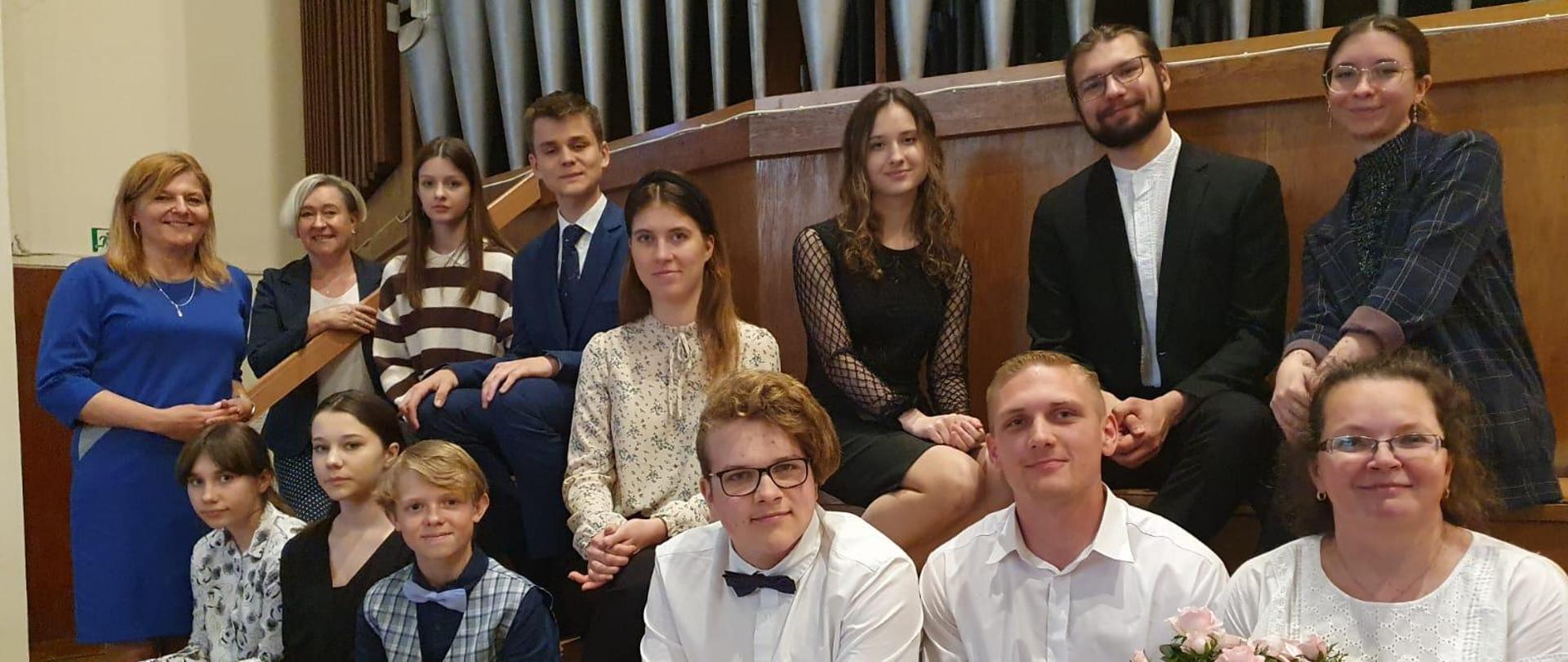 Na zdjęciu grupa młodych artystów, którzy wystąpili podczas koncertu w kościele parafialnym w Gdyni. W tle za nimi piszczałki organowej. W pierwszym rzędzie kobieta w okularach i białej bluzce, która trzyma różyczkę.