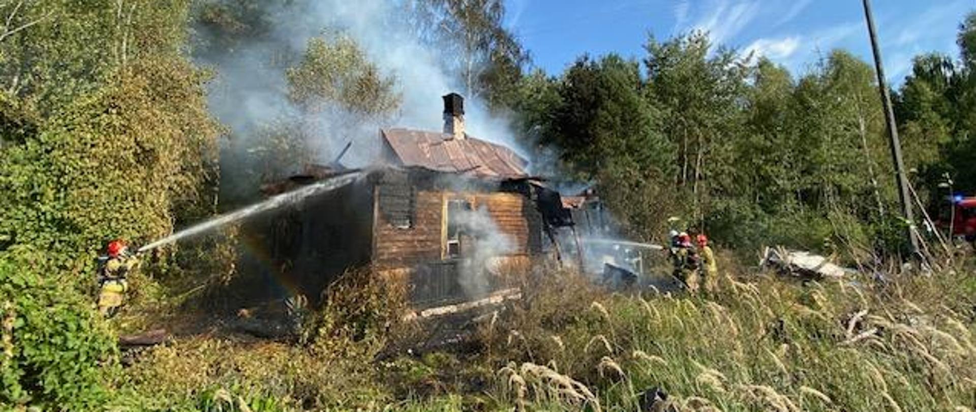 Na zdjęciu widoczny jest budynek mieszkalny jednorodzinny, konstrukcji drewnianej, który objęty był pożarem. Budynek z częściowo zniszczoną konstrukcją dachu. Na zdjęciu widoczni strażacy podający dwa prądy wody na palący się obiekt