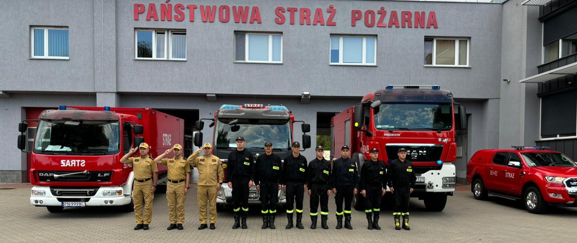 Strażacy stoją przed garażem i oddają hołd zmarłym strażakom z Poznania. Z tylu zaparkowane samochody strażackie na włączonych sygnałach świetlno-dźwiękowych. 