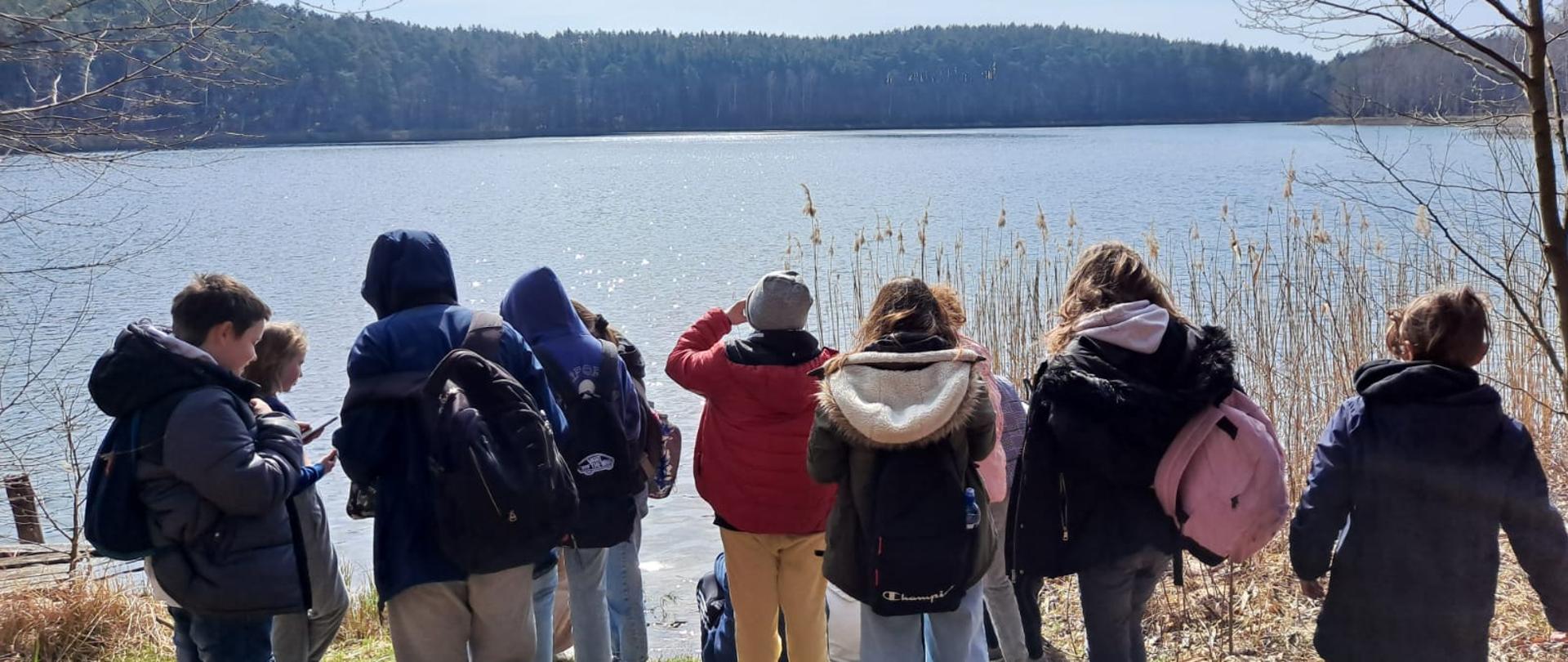 Grupa kilkorga dzieci stoi na ścieżce, na drugim planie jezioro.