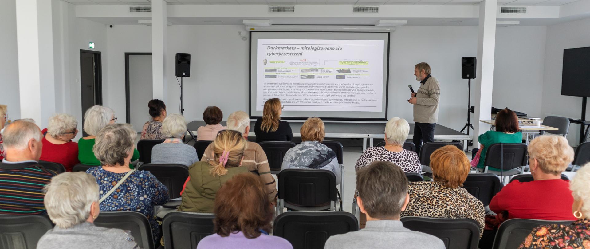 Grupa starszych osób siedzących na krzesłach w jasnym, przestronnym pomieszczeniu. Są skierowani w stronę mężczyzny, który stoi przed ekranem projekcyjnym i prowadzi prezentację. Ekran wyświetla slajd, na którym można dostrzec napis "Cyberprzestępczość". Mężczyzna trzyma w ręku mikrofon i przemawia do zebranych uczestników.