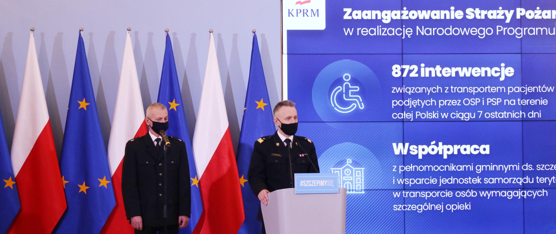Na zdjęciu dwóch strażaków w umundurowaniu wyjściowym jeden z OSP drugi z PSP. Obaj na twarzy mają maseczki. Za ich plecami ustawione są naprzemiennie flagi Polski i Unii Europejskiej. Obok ściana wizyjna, na której wyświetlane są informacje związane z konferencją.