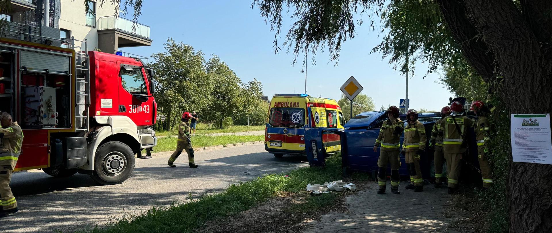 Po lewej stronie stoi samochód pożarniczy, dalej w prawo widać ambulans Zespołu Ratownictwa Medycznego, a skrajnie po lewej znajduje się leżący na boku samochód dostawczy wokół stoją ratownicy.