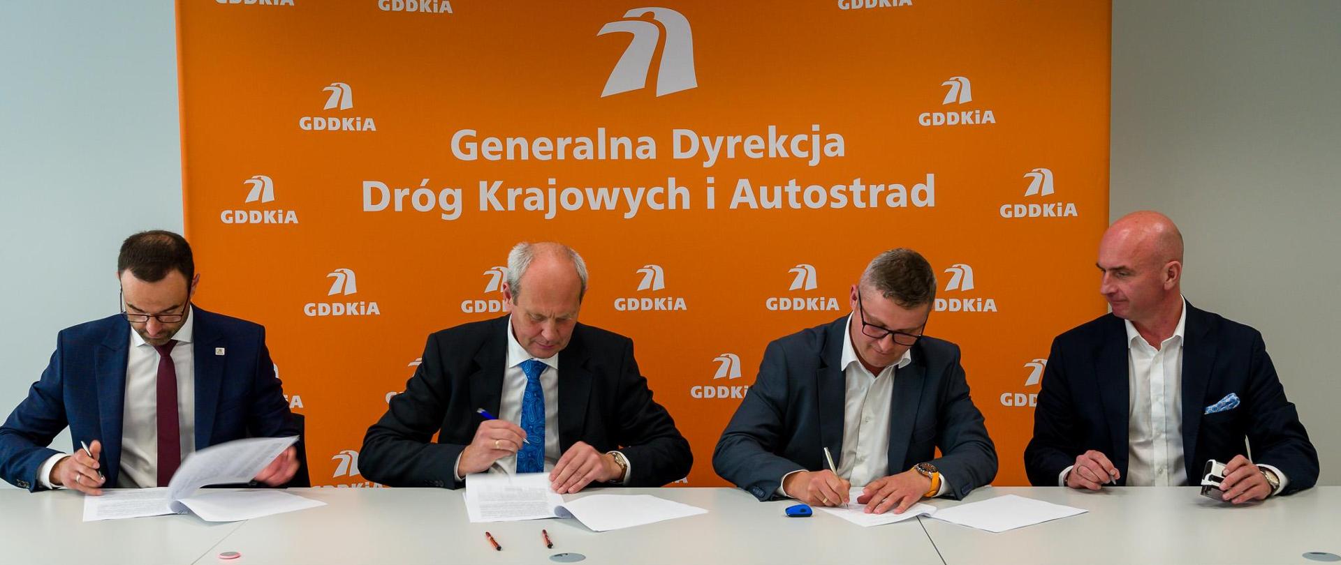 Moment podpisania jednego z 18 aneksów waloryzacyjnych. 4 elegancko ubranych mężczyzn siedzących obok siebie podpisuje dokumenty. W tle pomarańczowa ścianka z logotypami i nazwą GDDKiA.