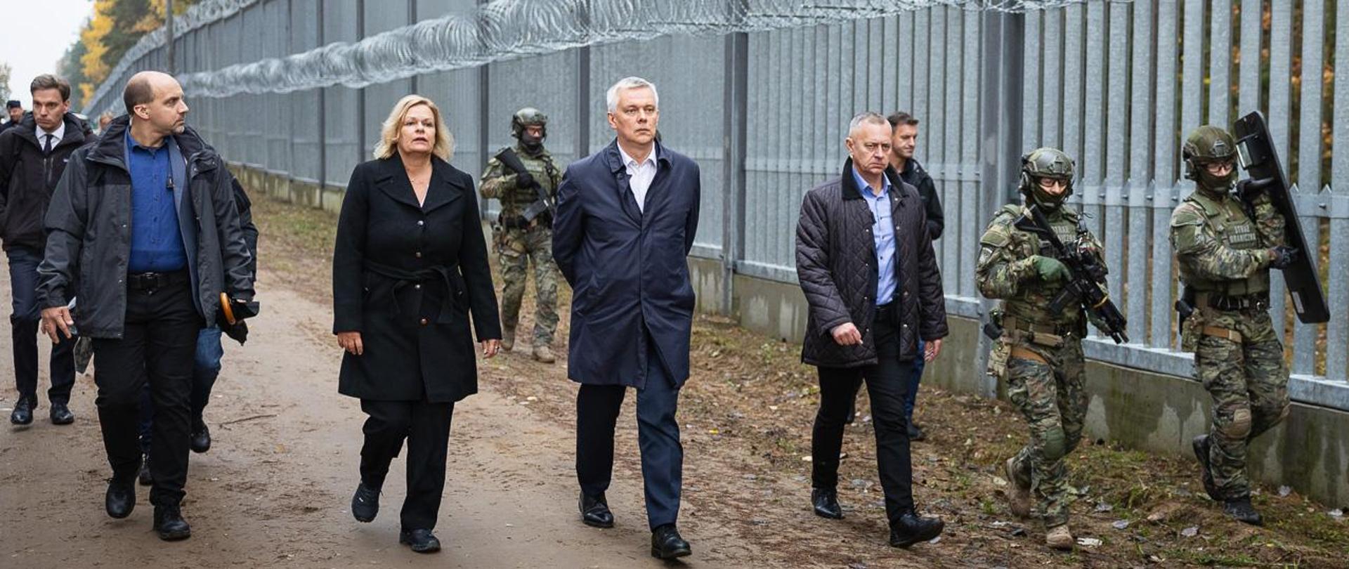 Ministrowie Tomasz Siemoniak i Nancy Faeser wizytują pas przy granicy polsko-białoruskiej. Towarzyszy im grupa ludzi. Zapory strzegą uzbrojeni strażnicy graniczni 