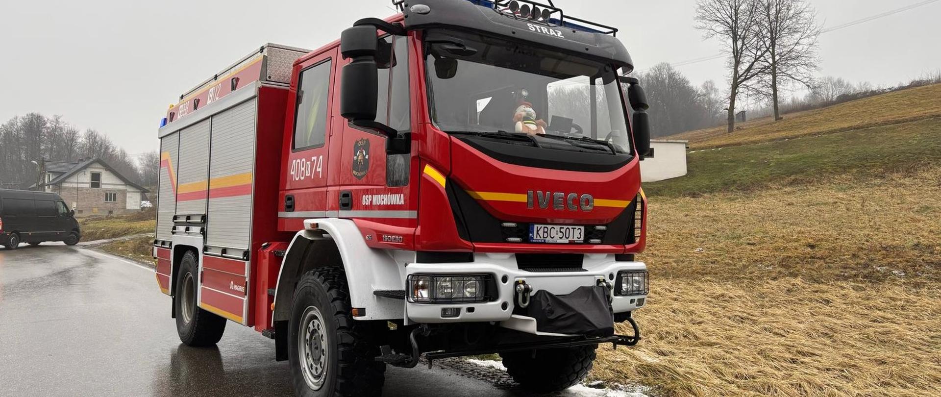 Na zdjęciu znajduje się samochód pożarniczy który zaparkowany jest na jezdni asfaltowej w tle widać łąkę, samochód typu bus i budynek mieszkalny 