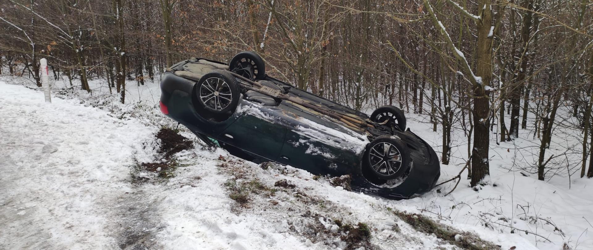 samochód osobowy leżący na dachu na poboczu drogi, widok z boku, na ziemi śnieg, w tle las