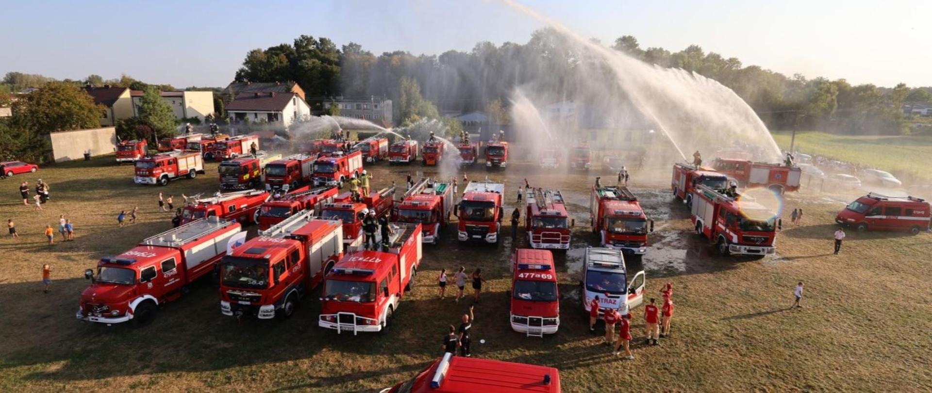 na trawistym placu znajduje się kilkadziesiąt samochodów pożarniczych z niektórych podawane są prądy wody, w oddali budynki i drzewa