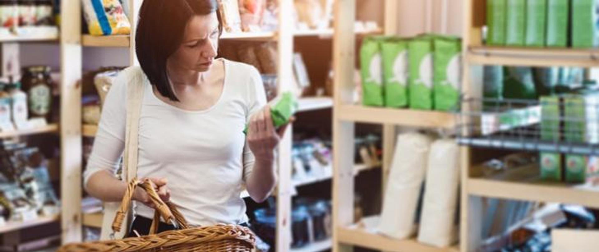 kobieta z koszykiem w ręku robi zakupy w sklepie. W ręku trzyma opakowanie i czyta opis produktu