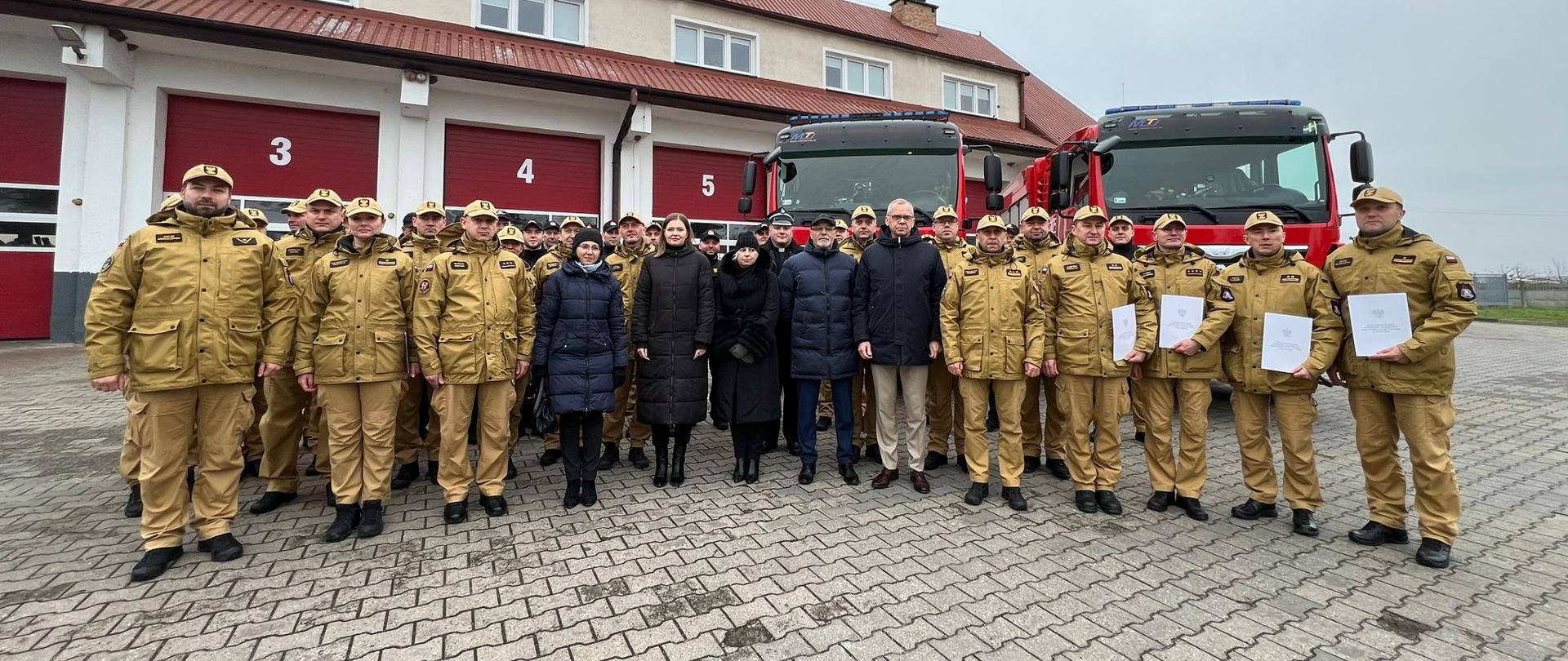 Wspólna fotografia funkcjonariuszy, przedstawicieli wojewódzkiego funduszu ochrony środowiska i gospodarki wodnej w szczecinie oraz samorządowców z powiatu pyrzyckiego, biorących udział w uroczystej zbiórce. W tle widoczny jest budynek komendy powiatowej psp w Pyrzycach oraz dwa czerwone samochody ratowniczo-gaśnicze