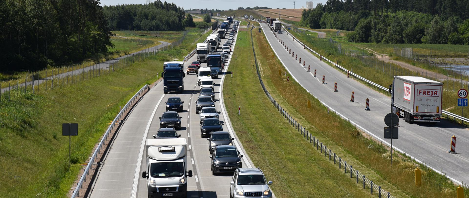 Droga z widocznym dużym zatorem po lewej stronie, gdzie sznur samochodów i ciężarówek porusza się wolno. Po prawej stronie drogi, oddzielonej pachołkami, ruch odbywa się płynnie, z jednym widocznym samochodem ciężarowym. Otoczenie drogi stanowią tereny zielone i drzewa, w tle także pola,