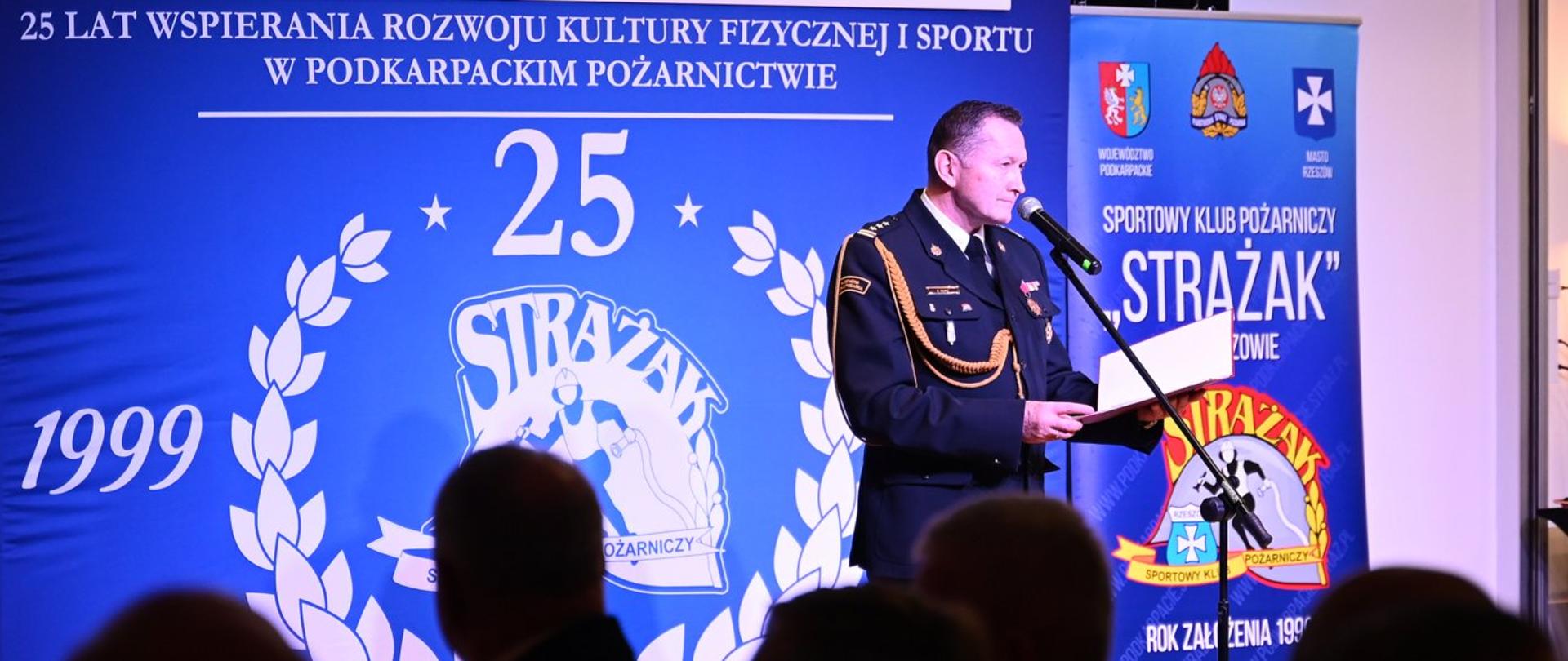 Zdjęcie zrobione podczas jubileuszowej gali z okazji 25-lecia działalności Sportowego Klubu Pożarniczego "Strażak" w Rzeszowie. Strażak w galowym mundurze przemawia do mikrofonu. W tle widać ekran z jubileuszowym logo "25 lat 1999-2024" oraz banery wydarzenia.