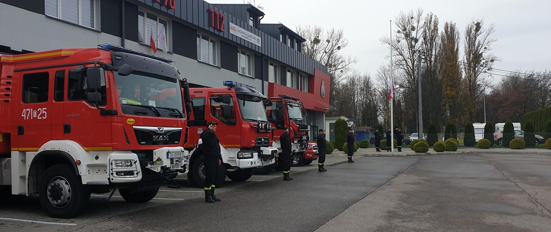 Zdjęcie przedstawia strażaków, którzy stoją przed samochodami strażackimi oddając honor podczas podnoszenia flagi państwowej.