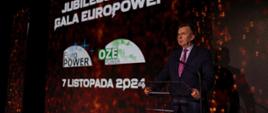 Na zaciemnionej sali na tle ściany w czarno-pomarańczowy wzorek z napisem Jubileuszowa gala EuroPower za przezroczystą mównicą stoi minister Wieczorek i mówi do mikrofonu.