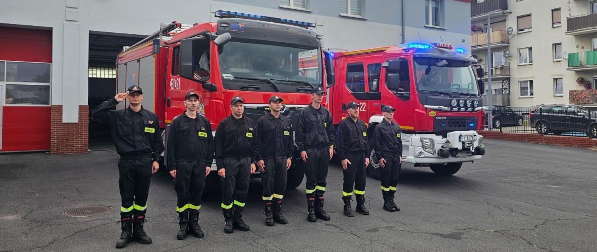 Strażaków oraz druhów stojących w szeregu przed wozami bojowymi. Jeden z nich oddaje honor poprzez salutowanie. Wozy mają włączone sygnały błyskowe.