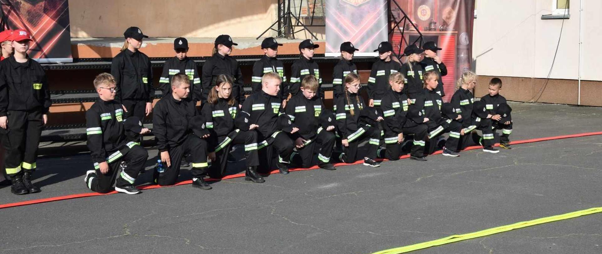 Przed sceną znajduje sie dziecięca drużyna pożarnicza. Członkowie drużyny ubrani są w mundurki koszarowe. Pierwszy szereg klęczy na czerwonym wężu, drugi - stoi. W składzie drużyny są zarówno dziewczęta jak i chłopcy.