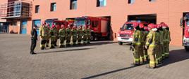 Uroczysta zmiana służby przed siedzibą JRG PSP Ostrów Wielkopolski. Strażacy stoją w czterech rzędach i oddają hołd.