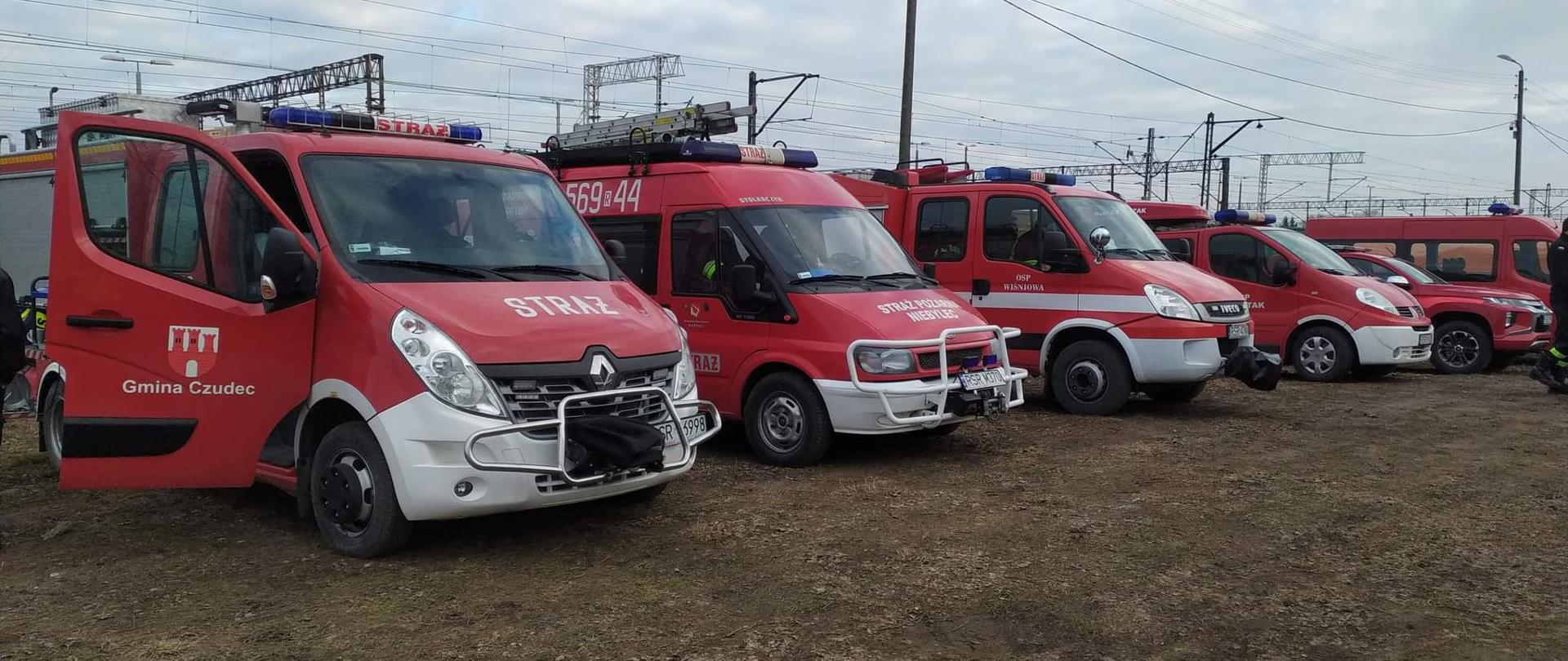 Na zdjęciu widoczne jest 5 samochodów strażackich jednostek z terenu powiatu Strzyżowskiego. Samochody w kolorze czerwonym stoją ustawione w szeregu. W tle widoczne linie trakcji kolejowej.