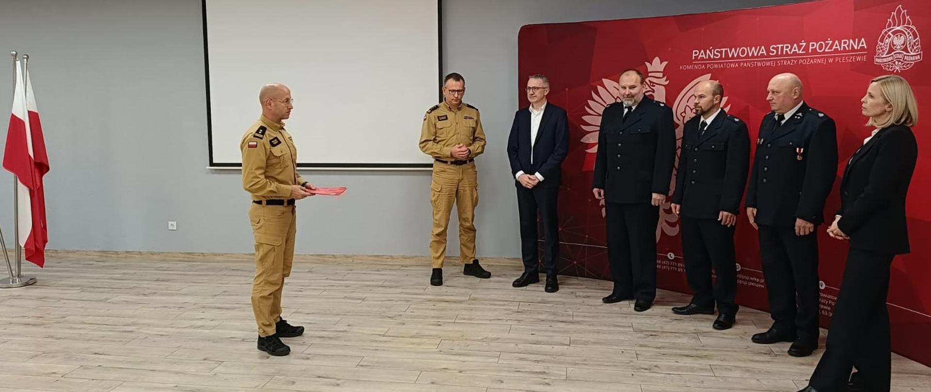 Na oświetlonej sali stoi w szeregu grupa mężczyzn i kobieta, trzech mężczyzn ubranych jest w mundury wyjściowe Ochotniczej Straży Pożarnej, jeden z mężczyzn ubrany jest w żółty mundur Państwowej Straży Pożarnej, obok niego stoi mężczyzna w ubraniu cywilnym, pierwsza z prawej stoi kobieta w ubraniu cywilnym, za nimi widoczna jest czerwona ścianka reklamowa z napisem Państwowa Straż Pożarna, na przeciw stojących w szeregu stoi mężczyzna ubrany w żółty mundur Państwowej Straży Pożarnej, i przemawia do zebranych