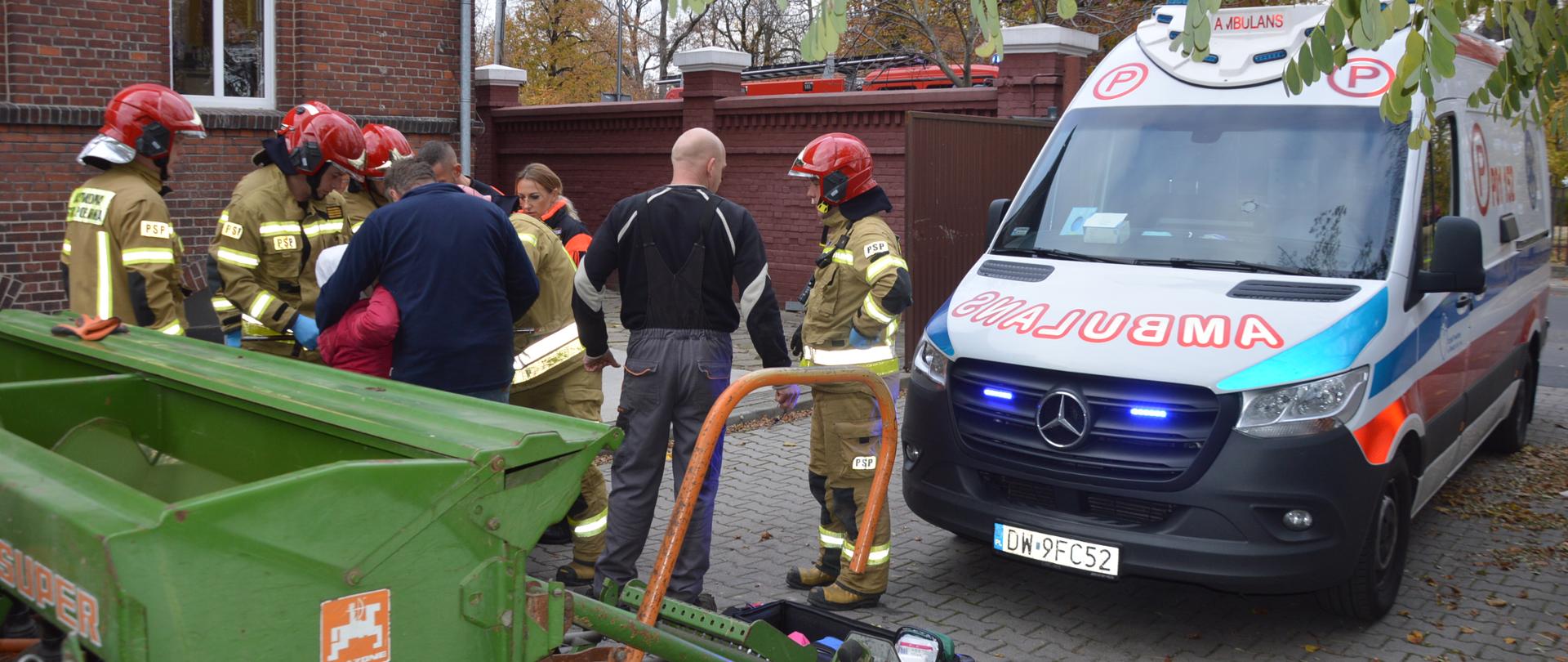 Wypadek podczas prac polowych. Teren jednego z zakładów pracy na terenie Rawicza. Strażacy i ratownicy medycznie udzielają pomocy kobiecie z urazem ręki. Z prawej strony karetka pogotowia ratunkowego. Na pierwszym planie fragment zielonego siewnika, a w tle budynek i mur z czerwonej cegły.