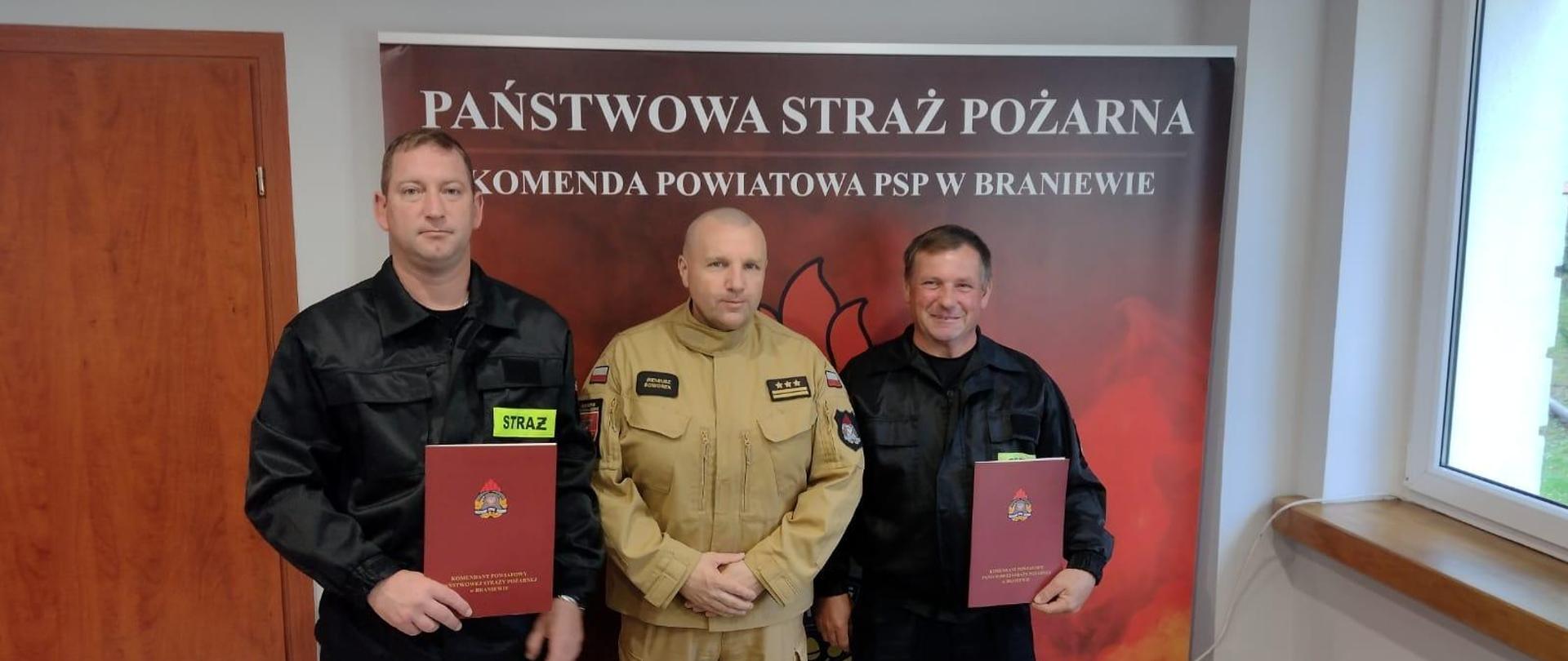 Na zdjęciu po środku stoi komendant powiatowy psp a po bokach dwóch strażaków ochotników. W tle czerwony baner Państwowa Straż Pożarna.