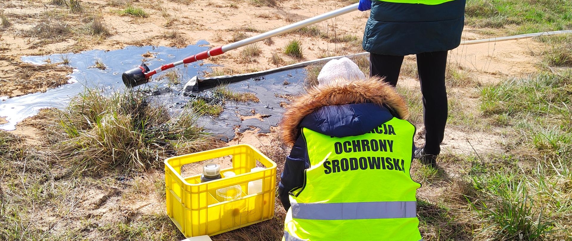 Dwie panie inspektor, w żółtych kamizelkach z napisem „Inspekcja Ochrony
Środowiska”, pobierają z powierzchni gruntu próbki wyciekającej cieczy.