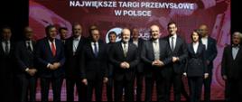 Na tle wielkiego czerwonego ekranu z napisem Największe targi przemysłowe w Polsce stoi grupa kilkunastu elegancko ubranych osób, pośrodku grupy wiceminister Gzik.