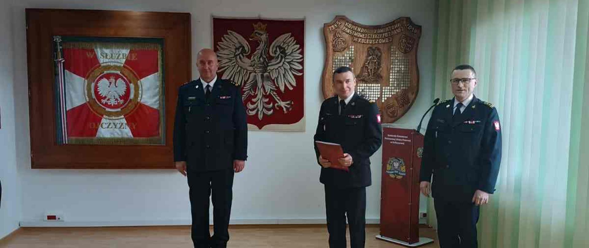 W pomieszczeniu stoi trzech strażaków w mundurach - jeden z nich trzyma czerwoną teczkę. W tle na ścianie sztandar i godło Polski.