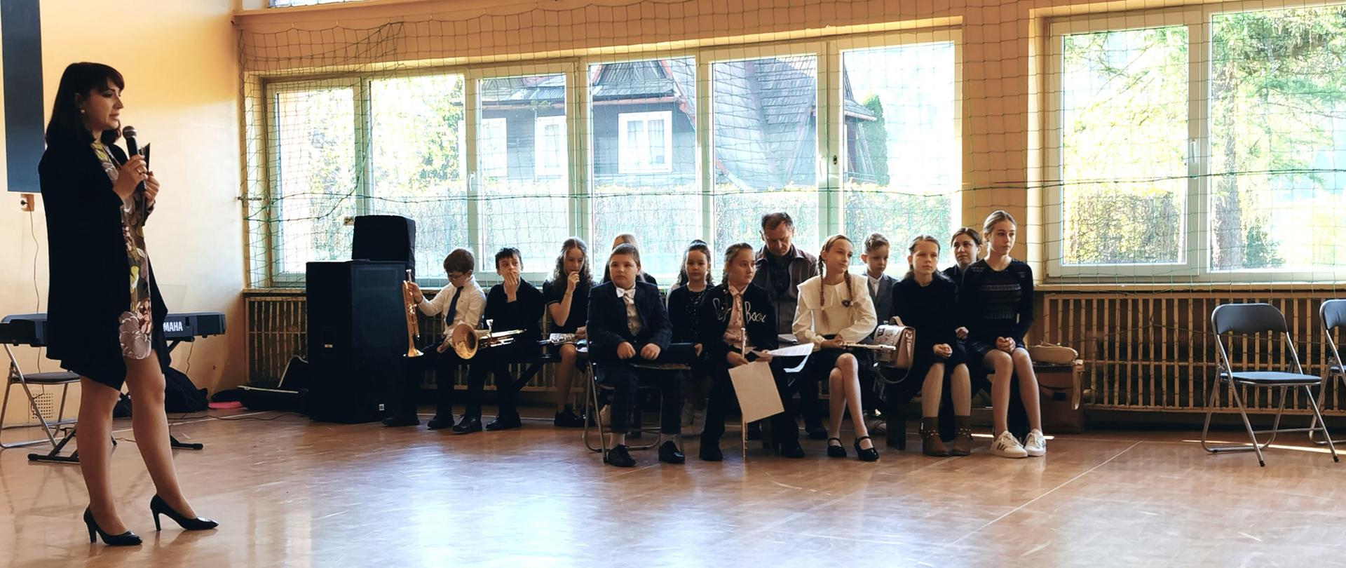 Sala gimnastyczna. Przed dziećmi nauczycielka szkoły muzycznej opowiada o zajęciach w szkole muzycznej, po prawej stronie uczniowie szkoły muzycznej siedzą na krzesłach w rękach trzymają instrumenty czekają na swój występ.